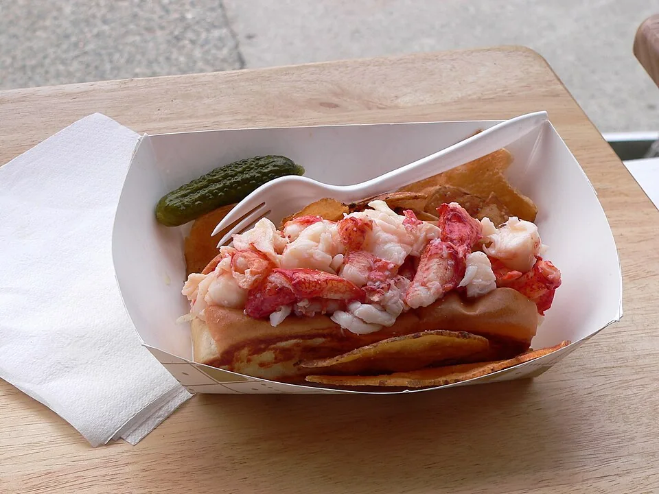 Lobster roll (estilo Maine): Bollo tostado en mantequilla relleno de carne fría de langosta ligada con mayonesa, apio y limón, ícono del verano de Maine.