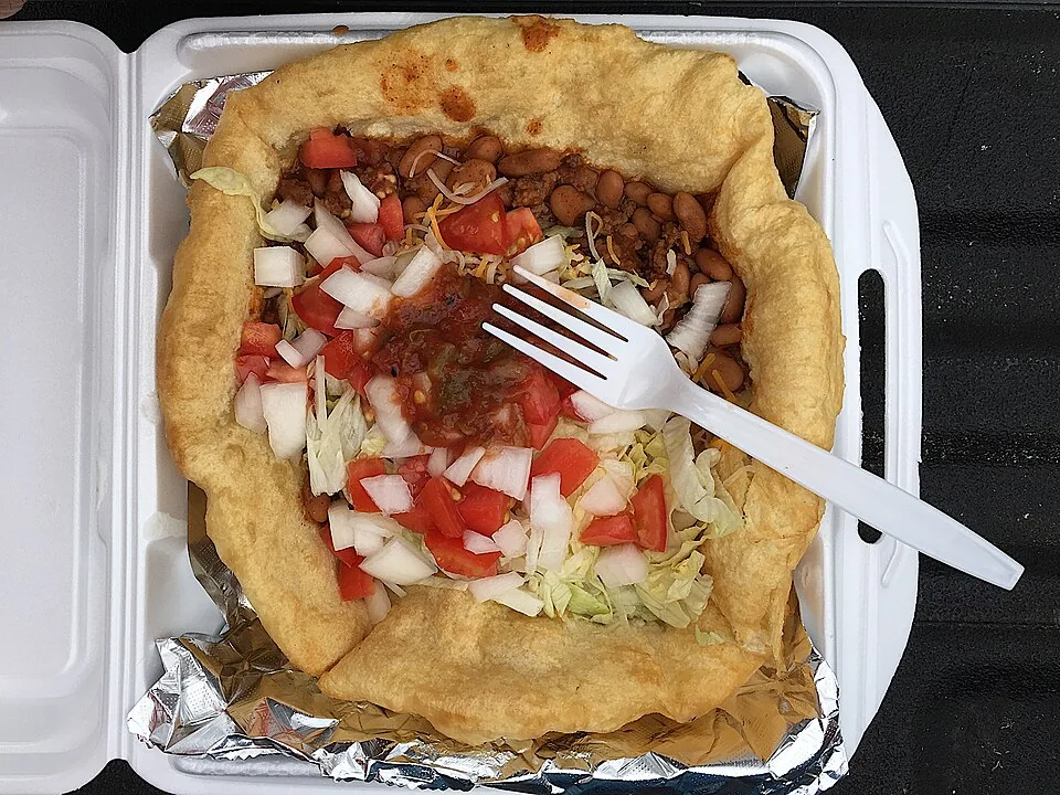 Navajo taco (fry bread): Pan frito esponjoso (fry bread) cubierto con frijoles, carne molida sazonada, lechuga, tomate y queso, servido como taco abierto.