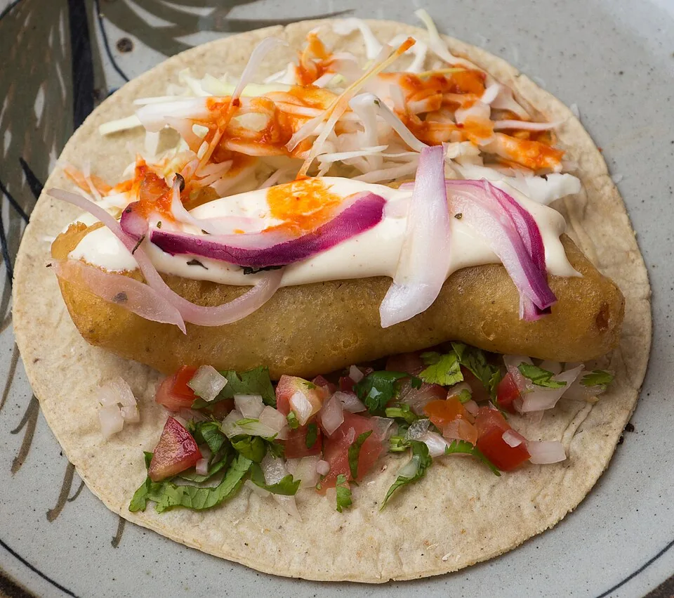 Tacos de pescado estilo Baja: Pescado capeado y frito en tortilla de maíz con col, crema, salsa y limón, sello de Ensenada.